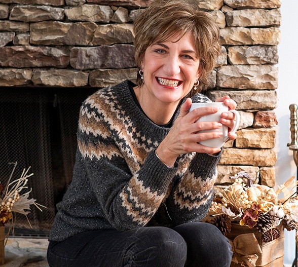 Karen Hardwick sitting by a fireplace smiling and holding a cup of coffee.
